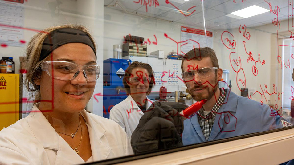 Students write out a chemical formula on a wipe board while the professor watches.